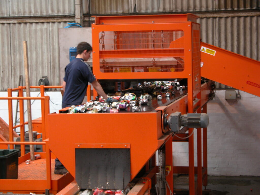 Image of workers hand-sorting materials at a can separation plant, with magnetic separation equipment visible in the background. data-large-image=