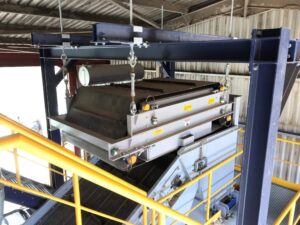 Image of a Magnapower electro overband magnet suspended above a conveyor belt, actively removing ferrous metals from a material stream.